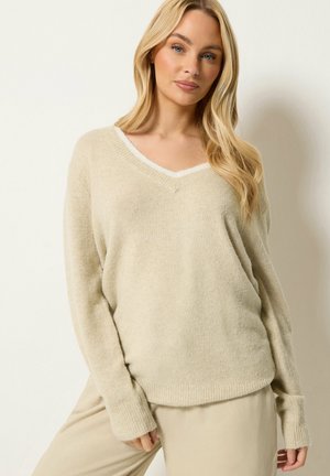 Kvinde med langt blondt hår iført en beige v-hals sweater og matchende bukser, står mod en ensfarvet lys baggrund.