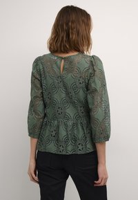 Zelená krajková blůza s tříčtvrtečními rukávy, květinovým vzorem, peplum detailem a klíčovou dírkou na zadním otevření. Texturovaný a jemný design.