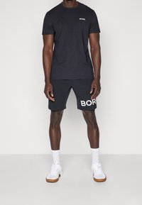 Schwarzes Kurzarm-T-Shirt mit weißem Logo, kombiniert mit schwarzen Shorts, die ein großes weißes Logo aufweisen. Getragen mit weißen Turnschuhen und Socken.