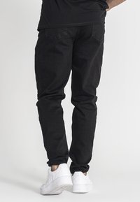 Pantalones de mezclilla negros con corte ajustado, que presentan bolsillos traseros y una textura suave, combinados con zapatillas blancas.