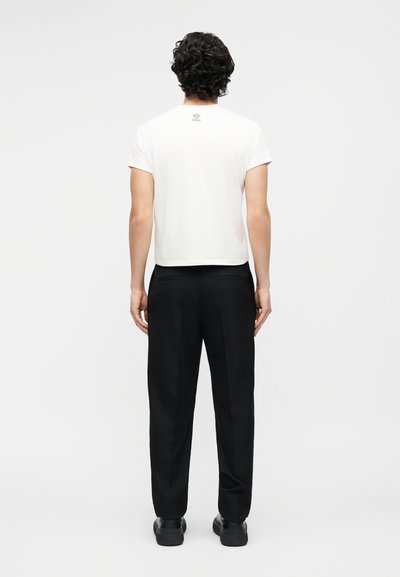 Άνδρας με σγουρά μαλλιά που φοράει λευκό κοντό T-shirt και μαύρο παντελόνι, όρθιος με την πλάτη προς το μέρος μας σε απλό ανοιχτό γκρι φόντο.