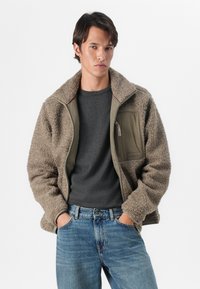 Lysebrun faux shearling jakke med en khaki lynlåslukning og lomme, båret over en mørkegrå skjorte, parret med blå jeans.
