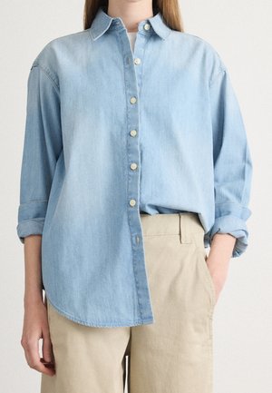 Person iført lys blå denimskjorte med knapper og ærmerne rullet op samt beige bukser, med den ene hånd i lommen, foran en ensfarvet baggrund.