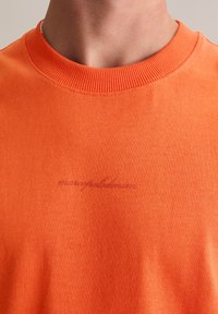 Pomarańczowy bawełniany t-shirt z prążkowanym dekoltem i subtelnym haftowanym logo w stylu kursywy. Gładka tekstura, prosty design bez wzorów.