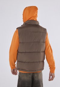 Ruda pūkinė liemenė su aukštu apykakle, dėvima virš oranžinės džemperio. Pasižymi quilted dizainu ir raukiniais kraštais. Atsitiktinis, sluoksniuotas apranga.
