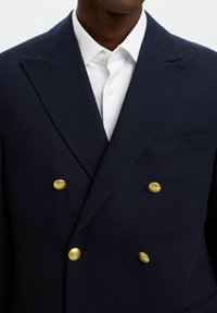 Granatowa marynarka na dwa rzędy guzików z teksturowanej tkaniny, ozdobiona złotymi guzikami i małą kieszonką na piersi. Do tego biała koszula zapinana na guziki.