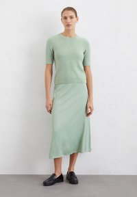 Maglione corto a maniche corte di un verde chiaro texturizzato abbinato a una gonna liscia di un verde chiaro di lunghezza media. La modella indossa mocassini neri.