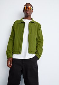 Green lightweight jacket with a zip closure, elastic cuffs, and two front pockets, worn over a white t-shirt and black pants, paired with reflective sunglasses.