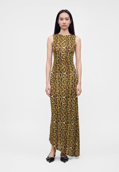 Femme aux longs cheveux noirs portant une robe sans manches, longue jusqu'au sol, jaune et marron à motif serpent, et des chaussures noires pointues.