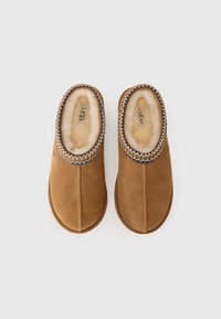 Un paio di pantofole in suede marrone con fodera in pile bianco e rifiniture decorative cucite, viste dall'alto su sfondo bianco.