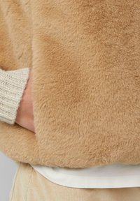 Hand in de zak van een zachte, tan kleurige neplederen jas, met een crèmekleurige gebreide mouw zichtbaar eronder.