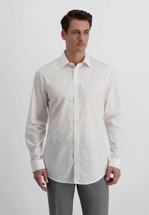 Man die een wit overhemd met lange mouwen en grijze broek draagt, staande tegen een eenvoudige lichte achtergrond.