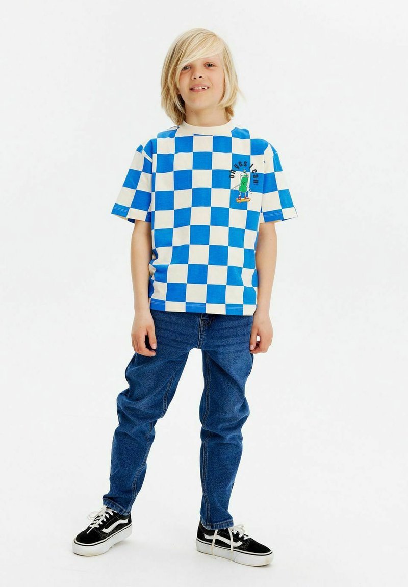 Camiseta de manga corta a cuadros azul y blanca con un gráfico en el pecho, combinada con jeans azules y zapatillas negras de corte bajo.