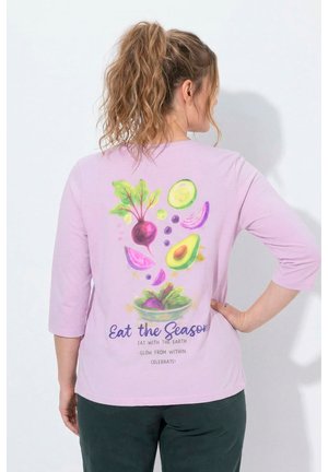 Vrouw met krullende paardenstaart die een lavendelkleurig shirt draagt en haar rug toont met kleurrijke groenteillustraties en tekst die seizoensgebonden eten promoot.