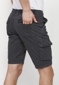 Man som bär mörkgrå cargo shorts med flera fickor, sätter handen i bakfickan, vit skjorta delvis synlig.