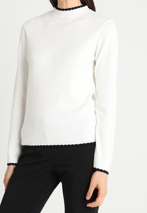 Femme portant un pull blanc à manches longues avec des bords festonnés noirs au col, aux poignets et à l'ourlet, associé à un pantalon noir.