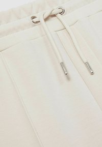 Pantaloni de training bej cu talie ajustabilă, cu ochiuri argintii și o textură moale, având un finisaj de material neted și fără modele.