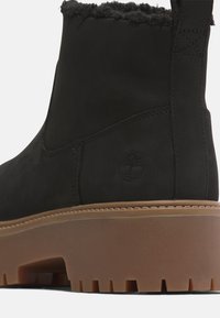 Black leather ankle boot with a soft fur lining around the collar, a rubber outsole featuring prominent tread, and a subtle logo embossed on the side.