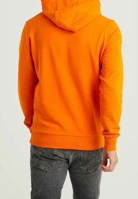 Persona che indossa un felpa con cappuccio arancione brillante e jeans neri sbiaditi, mostrata di spalle su uno sfondo chiaro e semplice.