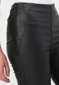 Pantalons taille haute en simili cuir noir avec une texture lisse, présentant une fermeture éclair sur le côté et des détails de couture le long des jambes.