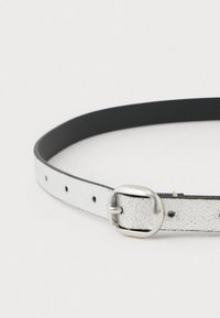 Sidabrinis įtrūkęs odinis diržas su lygiu juodu vidumi, turintis sidabrinį metalinį sagtį ir daugybę skylių reguliavimui.