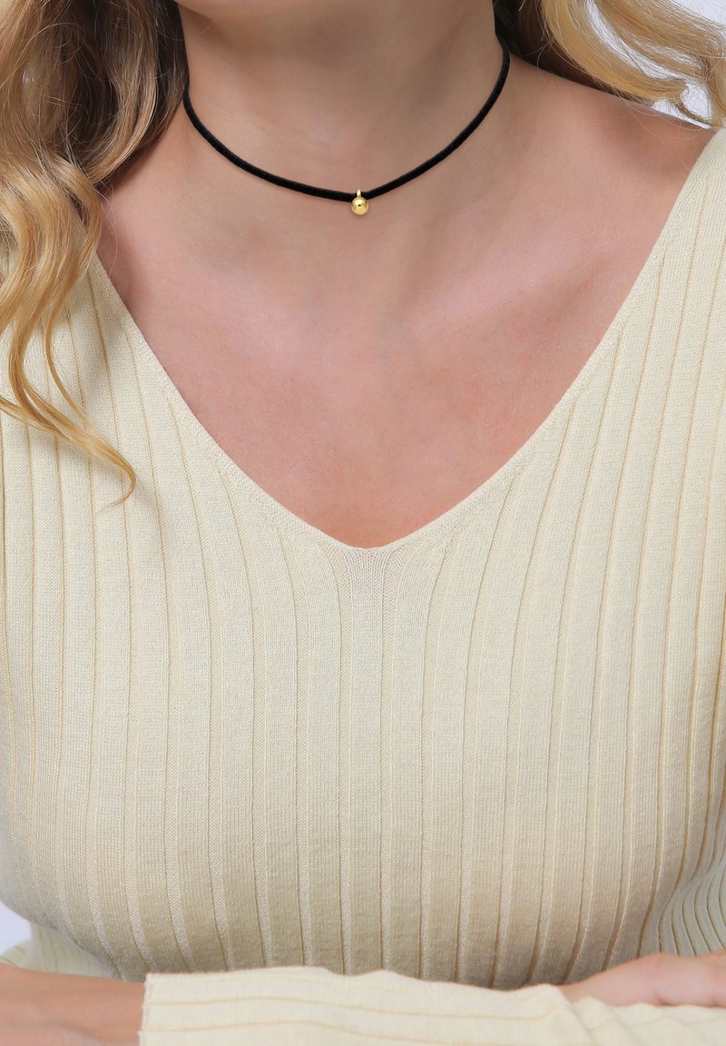 Collar choker negro hecho de cordón con un pequeño acento dorado en el centro. Llevado con un top de canalé en crema claro, que muestra un diseño de escote en V.