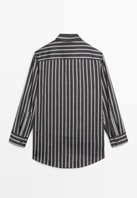 Chemise noire à manches longues avec des rayures blanches verticales et un yoke rayé horizontal dans le dos, posée à plat sur une surface blanche.
