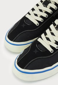 Black canvas sneakers with cream laces, blue accents along the sole, and star detail on the side. Rounded toe and textured fabric.