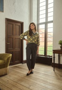 Femme en pull à motifs et pantalon foncé se tient près d'une grande fenêtre dans une pièce avec un sol en bois, un fauteuil vert vintage et une porte en bois.