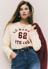 Polo crem cu mânecă lungă, având text brodat roșu "LE MARA 62 4TH ARR.", asociat cu blugi bleu din denim cu talie înaltă și o jachetă din blană burgundy.