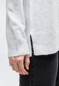 Χέρι με ασημένιο στριμμένο δαχτυλίδι ακουμπάει δίπλα σε μαύρο παντελόνι, φορώντας ανοιχτό γκρι πλεκτό πουλόβερ με ριμπ μανσέτες και πλευρικό σκίσιμο στο τελείωμα.