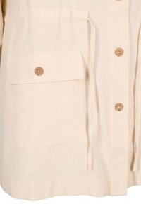 Vaaleahkot beige-nappijakku pehmeästä kankaasta, jossa on iso etutasku puunappilla ja säädettävä vyötärönauha.