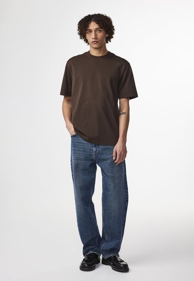 T-shirt in cotone marrone con scollatura a girocollo, abbinato a jeans in denim blu. Il modello indossa scarpe nere, mostrando una vestibilità rilassata.
