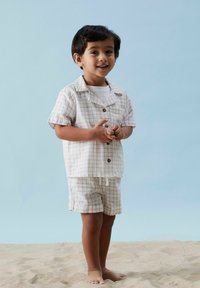 Lätt, beige rutmönstrad tvådelad outfit, kortärmad skjorta med knappar och matchande shorts, designad för komfort, bärs barfota på sand.