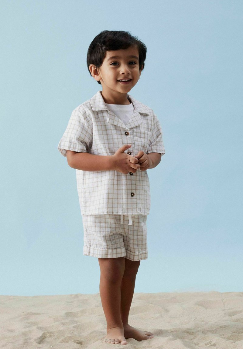 Lätt, beige rutmönstrad tvådelad outfit, kortärmad skjorta med knappar och matchande shorts, designad för komfort, bärs barfota på sand.
