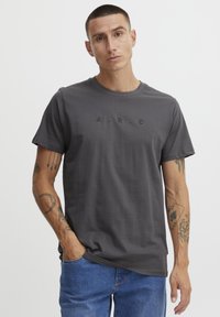 Mørk grå kortermet t-skjorte med teksten "A.R.C." trykt i svart. Glatt stoff, rund hals, løs passform, kombinert med lyseblå jeans.