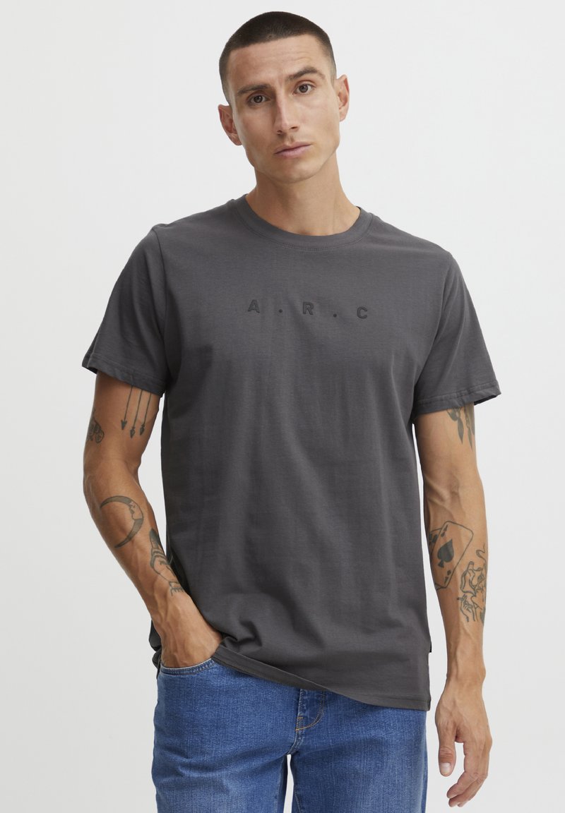Mørk grå kortermet t-skjorte med teksten "A.R.C." trykt i svart. Glatt stoff, rund hals, løs passform, kombinert med lyseblå jeans.
