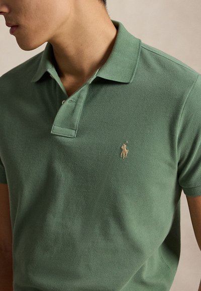 Vyras dėvi žalią polo marškinėlį trumpomis rankovėmis su mažyčiu smėlio spalvos arklio ir raitelio siuvinėjimu ant krūtinės.