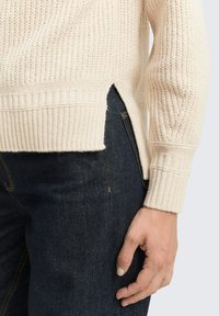 Beige gebreide trui met ribbels aan de zoom en mouwen, met zijsplitsen. Gepaard met donkerdenim jeans, die textuur en stofdetails tonen.