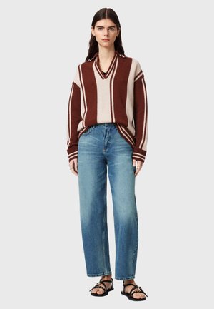Kvinde med langt mørkt hår iført brun og beige stribet sweater, løse blå jeans og sorte sandaler, står mod en ensfarvet baggrund.