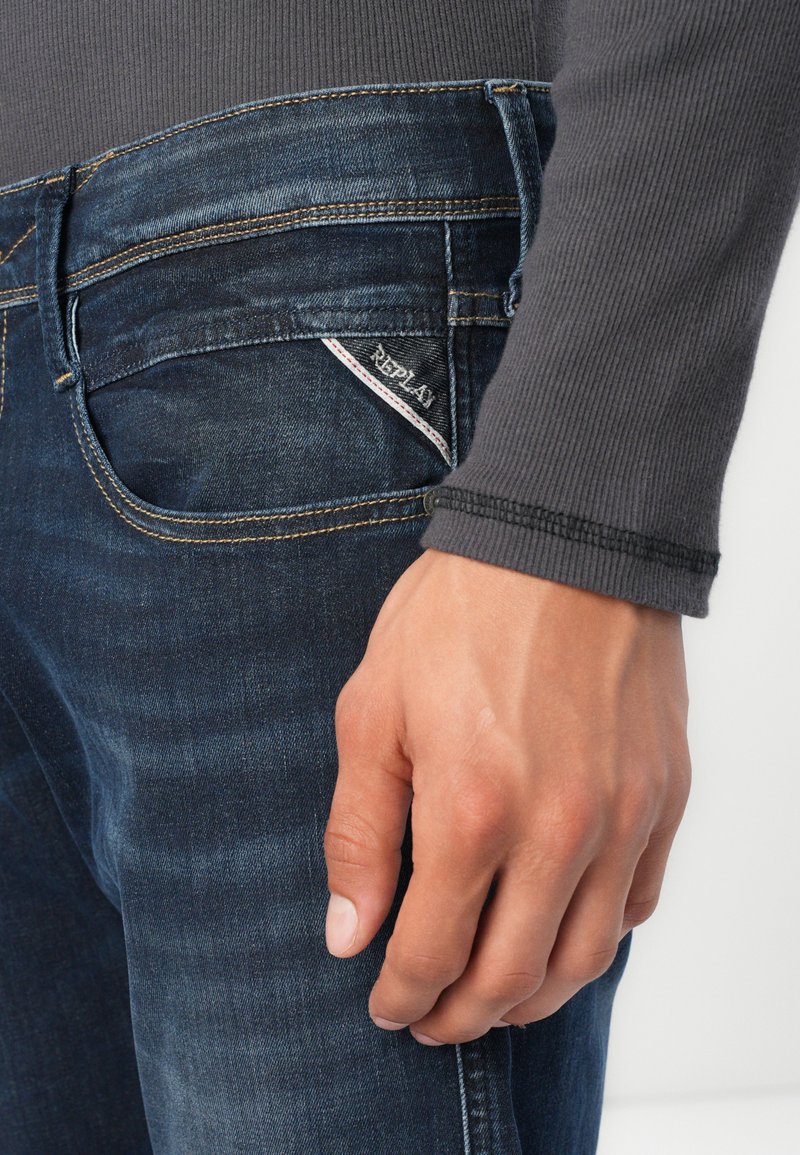 Dunkelblaue Jeans aus Denim mit auffälligen Nähten, strukturierter Oberfläche und kleinem "Replay"-Logo auf der Gesäßtasche. Gezeigtes graues Langarmoberteil.
