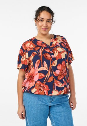 Woman wearing a navy blue short-sleeve floral blouse with pink, red, and beige flowers and light blue jeans, smiling at the camera.