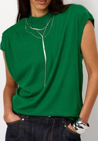 Groene top met korte mouwen, een hoge hals en een gedrapeerd ontwerp; beschikt over een zilveren ketting en een glanzende zilveren armband.