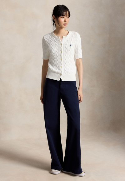 Kobieta z czarnymi włosami związanymi nisko w kucyk, ubrana w biały krótki sweter na guziki z grubym splotem, granatowe szerokie spodnie i białe tenisówki, stojąca.