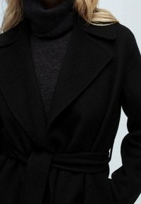 Manteau portefeuille noir en tissu doux, avec un grand col, une ceinture à la taille et des manches longues. En dessous, un col roulé sombre est visible.