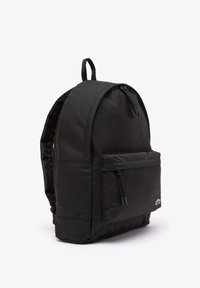Mochila negra hecha de tela resistente, con una parte superior redondeada, un bolsillo frontal con cremallera y correas de hombro ajustables. Acento del logo cerca de la base.