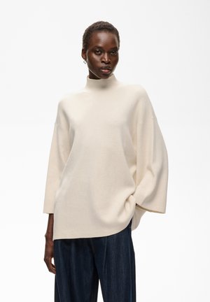 Femme portant un pull col roulé ample de couleur crème avec de larges manches et un pantalon large bleu foncé sur un fond blanc uni.