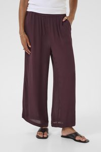 Woman wearing loose, wide-leg dark purple pants with an elastic waistband, white top, and black open-toe sandals on a plain background.