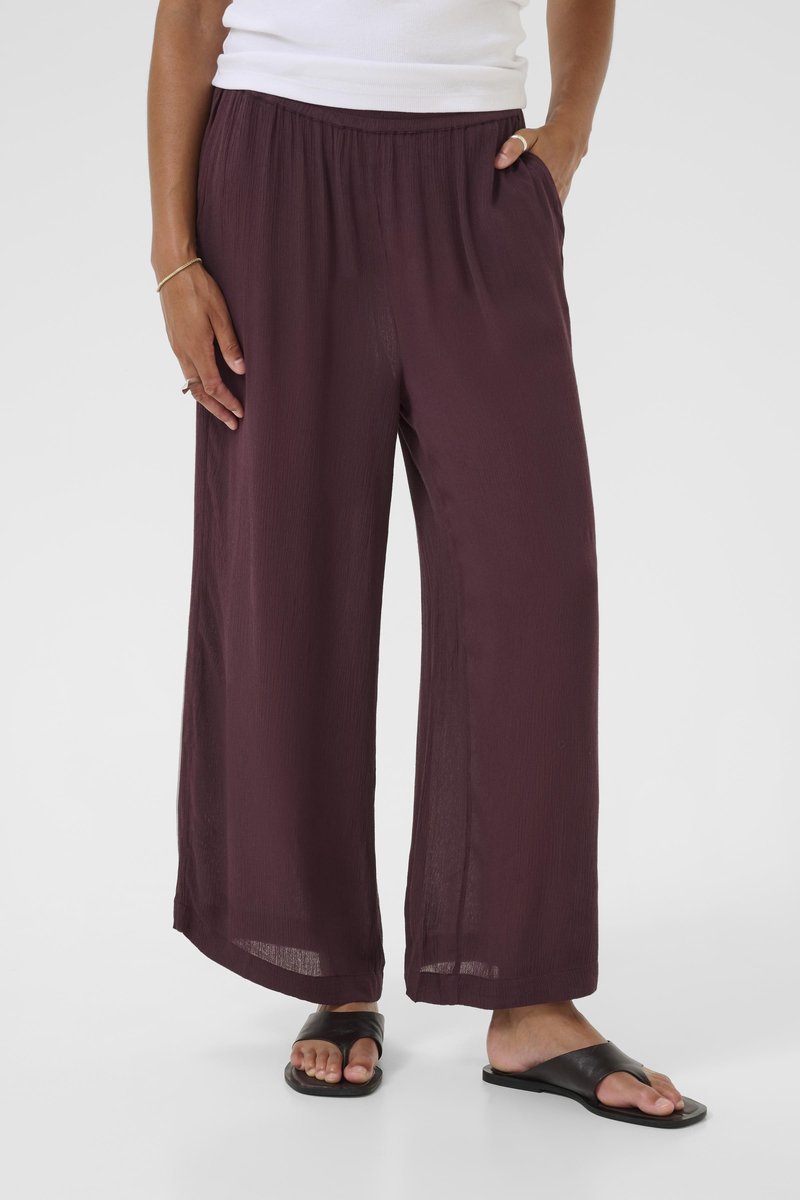 Woman wearing loose, wide-leg dark purple pants with an elastic waistband, white top, and black open-toe sandals on a plain background.
