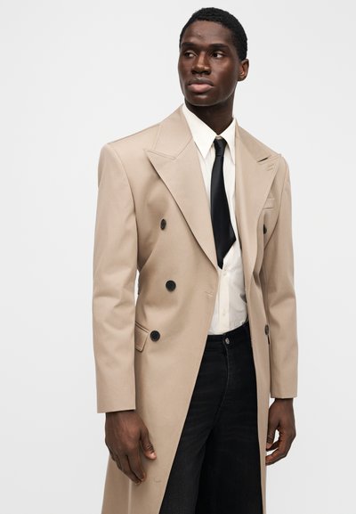 Abrigo beige de doble botonadura con grandes botones negros, solapas pico y hombros estructurados, combinado con una camisa blanca y corbata negra, llevado sobre vaqueros oscuros.